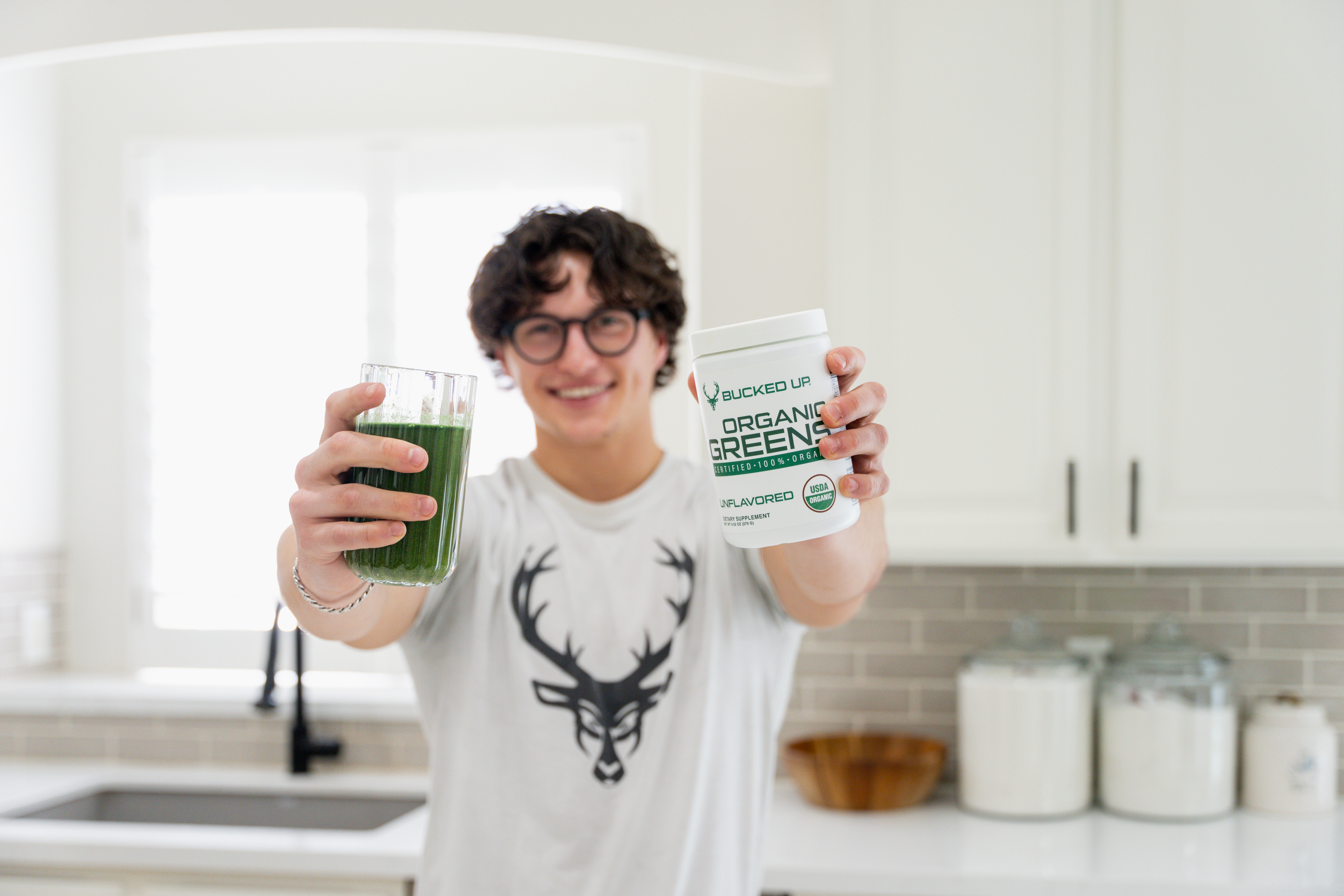 Smiling individual holding Bucked Up Organic Greens supplement container labeled 'USDA Organic, Unflavored,' and a glass of green drink in a bright, modern kitchen.