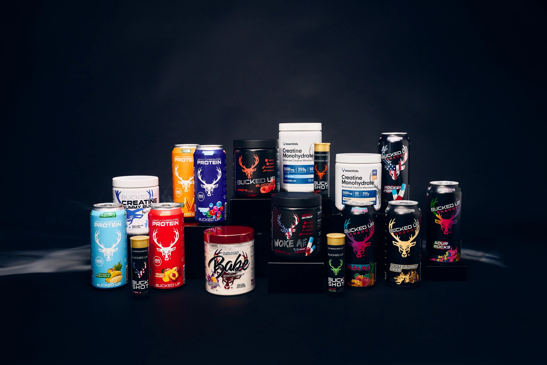 A collection of dietary supplement containers and energy drink cans arranged on a black surface against a dark background, with some containers stacked and others standing upright, featuring vibrant colors and bold branding, including logos of a deer head and energetic graphics, illuminated by soft, focused lighting that highlights the labels and textures of the packaging, creating a professional and dynamic presentation.
