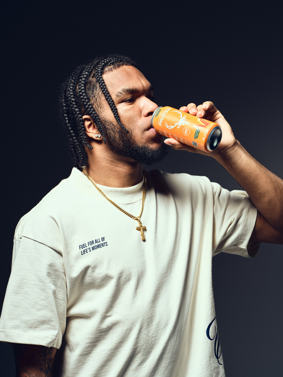 A young man with braided hair, a beard, and earrings is drinking from an orange can with a deer logo, wearing a beige T-shirt with the phrase "Fuel for all of life's moments" and a gold chain with a cross pendant, against a dark background, captured in a professional studio setting with soft lighting highlighting his profile and the can.