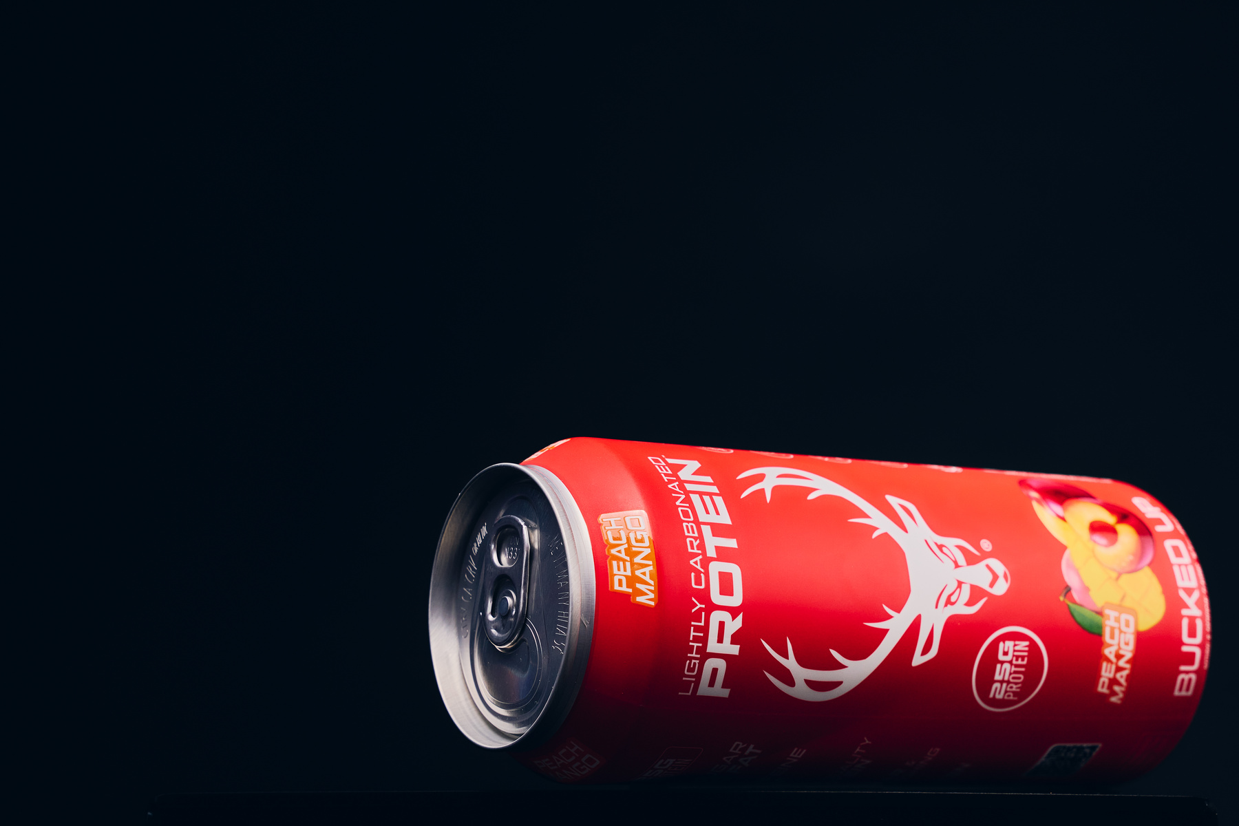 A red aluminum can of Bucked Up Protein Soda drink lies on a black reflective surface against a dark background, positioned horizontally with the pull-tab facing the viewer. The can features a white stylized deer head logo, the words 'PEACH MANGO' in orange and white, and 'LIGHTLY CARBONATED' and '25g PROTEIN' in smaller white text. The design includes a vibrant illustration of a peach and mango fruit at the bottom right. The lighting highlights the can's glossy surface, creating subtle reflections and shadows that emphasize its cylindrical shape, with a professional studio style and a sleek, energetic mood.