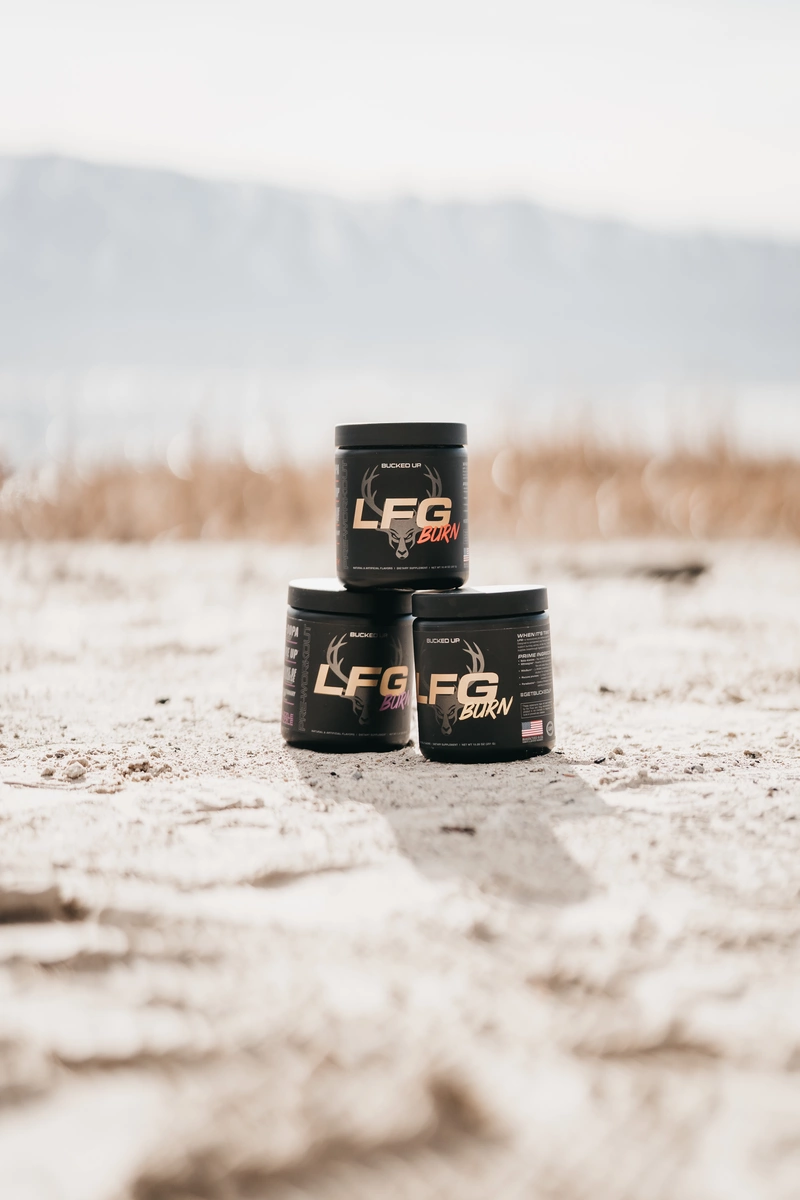 Three black containers of LFG BURN supplement with a deer logo and bold white and red text are stacked on sandy ground outdoors, with a blurred background of dry grass, distant mountains, and a clear sky, captured from a low angle in natural daylight, emphasizing the product's branding and rugged outdoor setting.