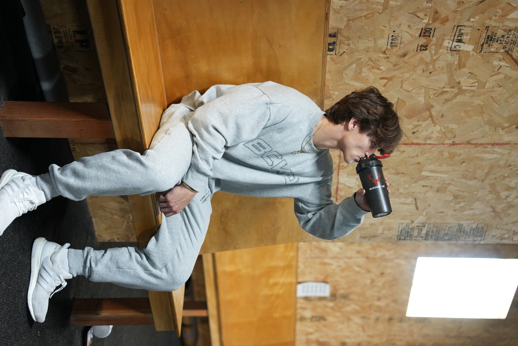 A young man with wavy brown hair and light skin, dressed in a light gray oversized sweatshirt with a subtle logo, matching sweatpants, and white athletic shoes, is sitting on a wooden bench in a room with unfinished plywood walls. He is drinking from a black water bottle with a red logo, holding it with his right hand, while his left hand rests on his lap. The room has a high window allowing bright natural light to illuminate the scene, creating soft shadows. The background includes a small white device on the wall and some printed markings on the plywood, with a casual, relaxed atmosphere and a focus on comfort and simplicity.