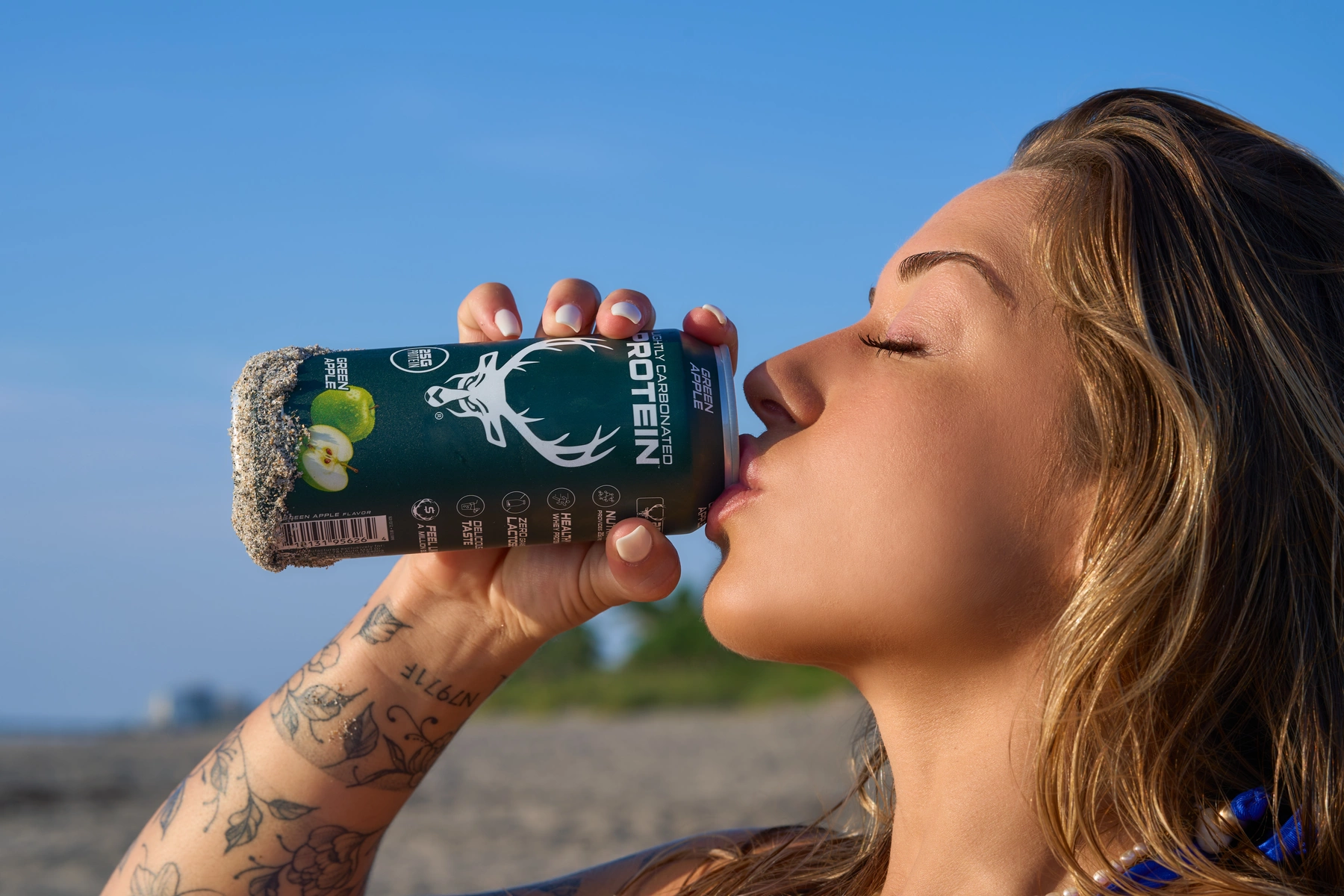 A young woman with wavy blonde hair, closed eyes, and a serene expression is drinking from a black can of Green Apple flavored protein drink with a sandy rim, held in her right hand, which has floral tattoos. She is outdoors on a sandy beach with a clear blue sky in the background, and a distant building is visible. The lighting is bright and natural, highlighting her smooth skin and the details of the can, with a relaxed, summery mood.