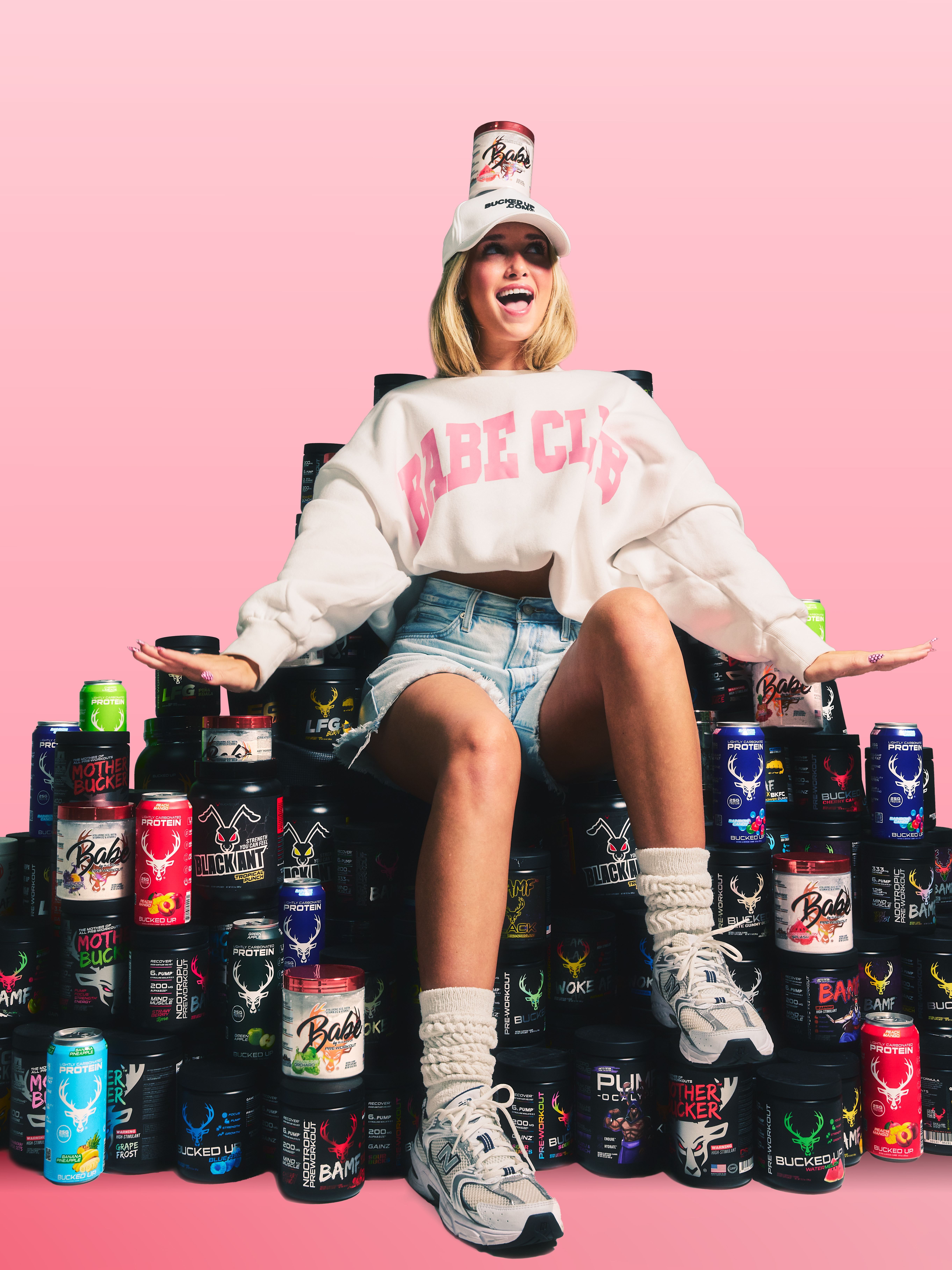 Smiling woman wearing Babe Club sweatshirt and Bucked Up hat sitting on stack of Bucked Up supplements with Babe protein tub on head – fun fitness lifestyle and supplement brand photo.