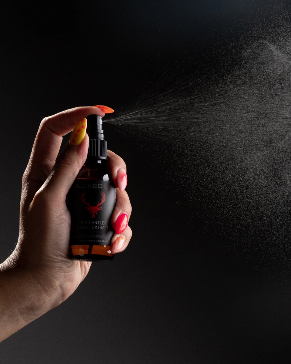 A close-up of a hand with manicured nails holding a small spray bottle labeled 'Deer Antler Velvet Extract' with a red deer head logo, spraying a fine mist against a dark, gradient background. The hand is positioned in the lower left, with the spray directed towards the upper right, capturing the spray particles in mid-air illuminated by soft lighting that highlights the skin and nails.