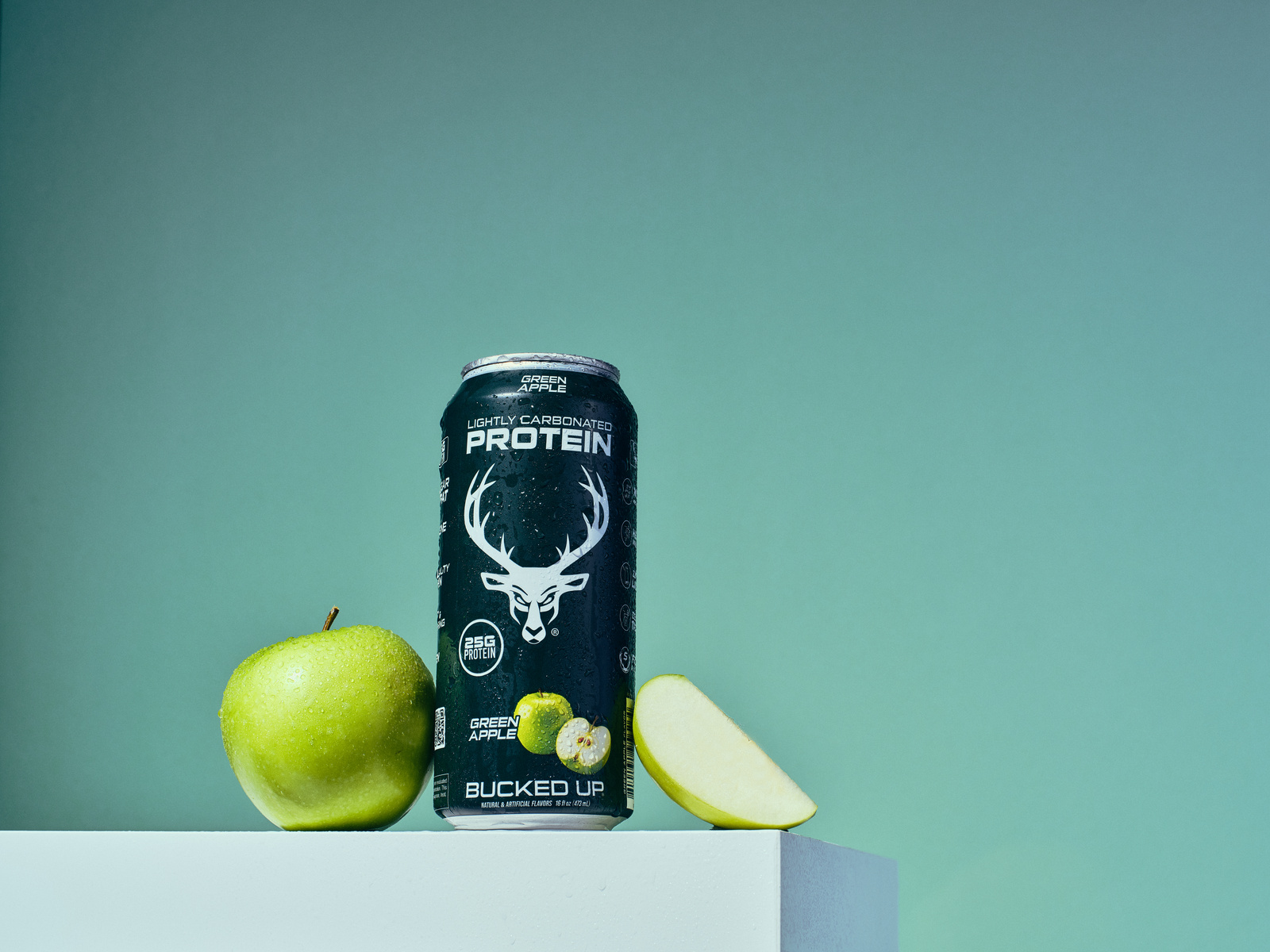 A can of Green Apple flavored protein drink with a sleek black design featuring a white deer head logo, placed on a white surface alongside a whole green apple with water droplets and a sliced wedge of the apple, set against a gradient green background. The composition is minimalistic with soft lighting highlighting the freshness of the apple and the modern design of the can, evoking a healthy, energetic mood.