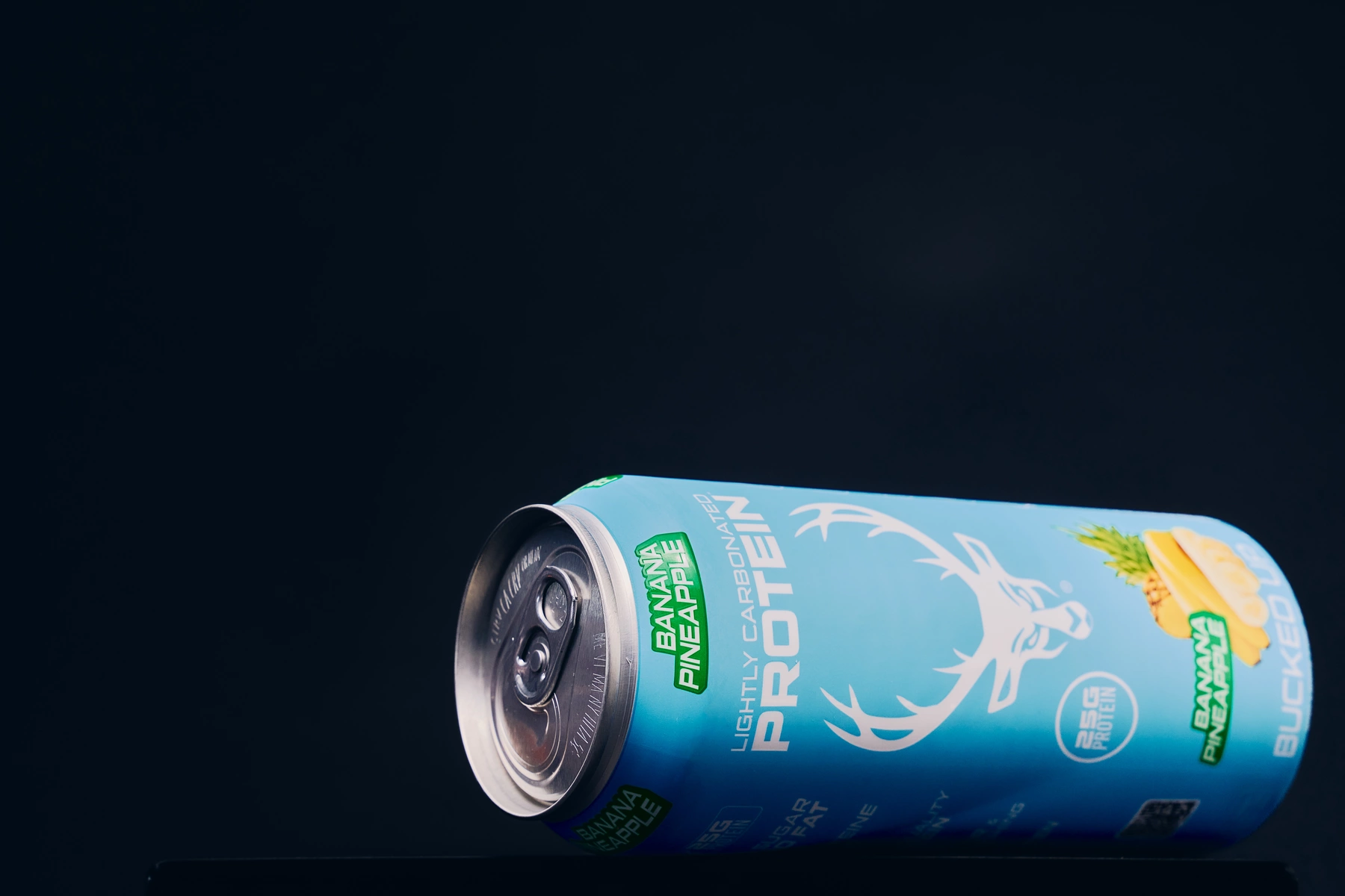 A light blue can of ProTein lightly carbonated beverage with a banana and pineapple flavor, featuring a white stylized deer head logo and green accents, lying on its side against a dark, almost black background with subtle lighting highlighting the metallic top and vibrant label details.