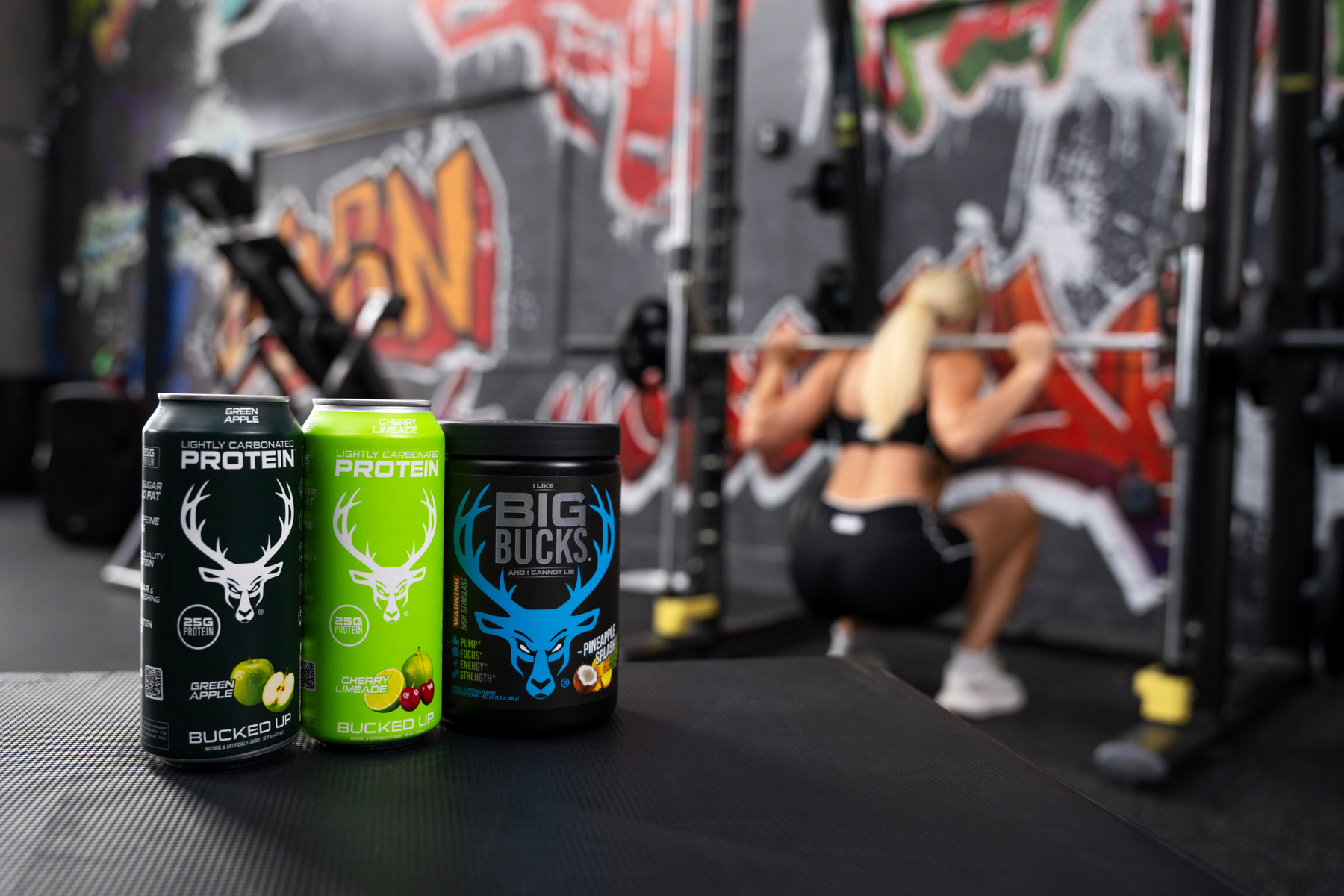 Three cans of protein drinks with black, green, and black labels featuring a white deer head logo are placed on a black gym bench in the foreground. The cans are labeled 'Lightly Carbonated Protein' with flavors 'Green Apple,' 'Cherry Limeade,' and 'Juiced.' In the background, a blonde woman with a ponytail, wearing a black sports bra and black shorts, is squatting with a barbell on her shoulders in front of a graffiti-covered wall. The gym environment includes black weight racks, yellow weights, and a dark, gritty atmosphere with colorful graffiti art on the wall. The lighting is bright and focused on the cans, with softer ambient light illuminating the woman and gym space.