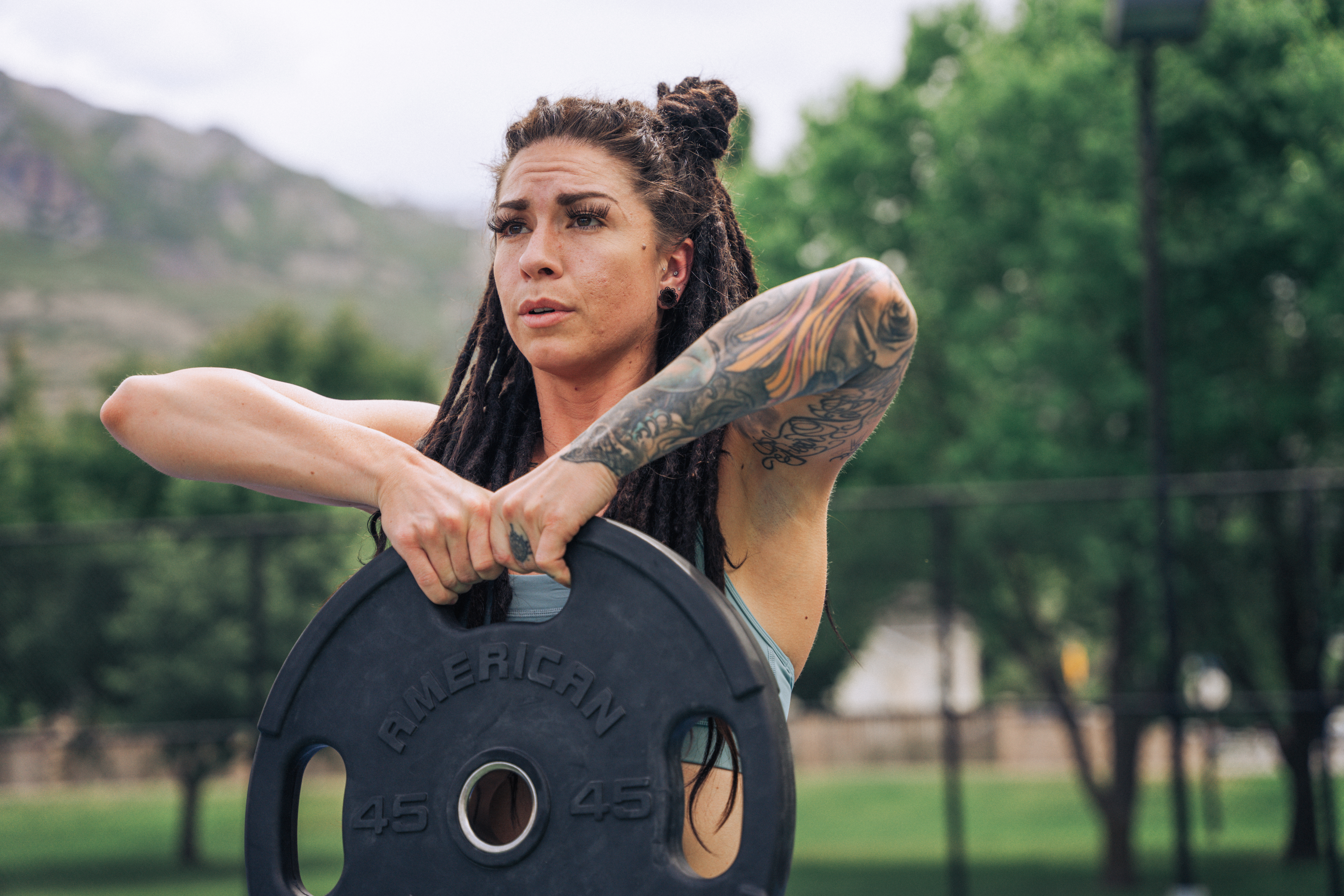 A woman with long, dark dreadlocks styled in a bun, wearing a light blue sports top, is outdoors in a park with lush green trees and mountains in the background. She has multiple tattoos on her right arm, including colorful designs and script, and is holding a black 45-pound weight plate labeled 'American' with both hands, performing a workout. Her facial expression shows focus and determination, with natural lighting highlighting her features and the vibrant colors of her tattoos. The scene captures a professional, energetic fitness moment with a natural, scenic environment.