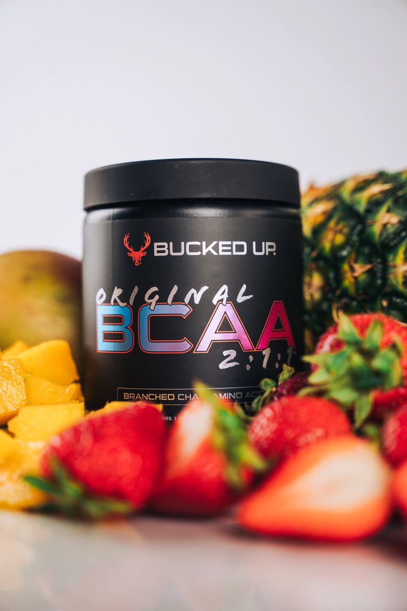 A black container of Bucked Up Original BCAA supplement with a red deer logo and colorful text, placed among fresh strawberries, pineapple chunks, and a pineapple in the background, on a white surface with a plain light gray background, shot from a slightly low angle with soft lighting highlighting the product and fruits, creating a vibrant and energetic mood.