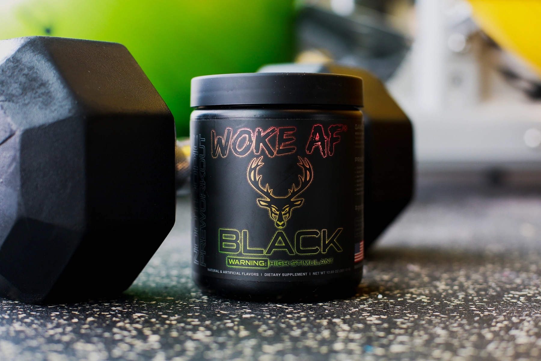 A black container of dietary supplement labeled 'WOKE AF BLACK' with a stylized deer head logo, placed on a textured black surface with scattered white specks, surrounded by a gray hexagonal dumbbell on the left and blurred gym equipment in the background, illuminated by natural light creating subtle highlights and shadows, with a vibrant green and yellow background.

