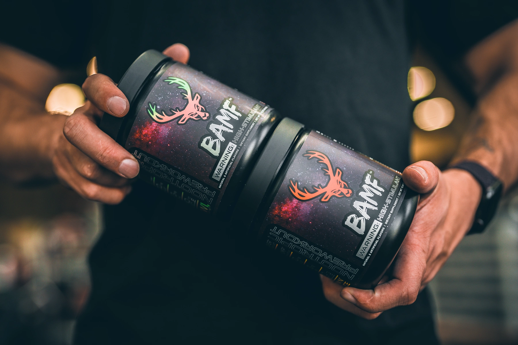 A close-up shot of two black containers of a pre-workout supplement labeled 'BAMF,' featuring a vibrant galaxy-themed design with a prominent orange deer logo and bold white text. The containers are held in a person's hands, showcasing muscular arms with a watch on the wrist. The background is softly blurred with warm bokeh lights, suggesting a gym environment. The lighting is dramatic, highlighting the containers and casting subtle shadows, creating a dynamic and energetic mood. The overall color palette includes deep blacks, bright oranges, and cosmic purples, emphasizing a modern and intense aesthetic.

