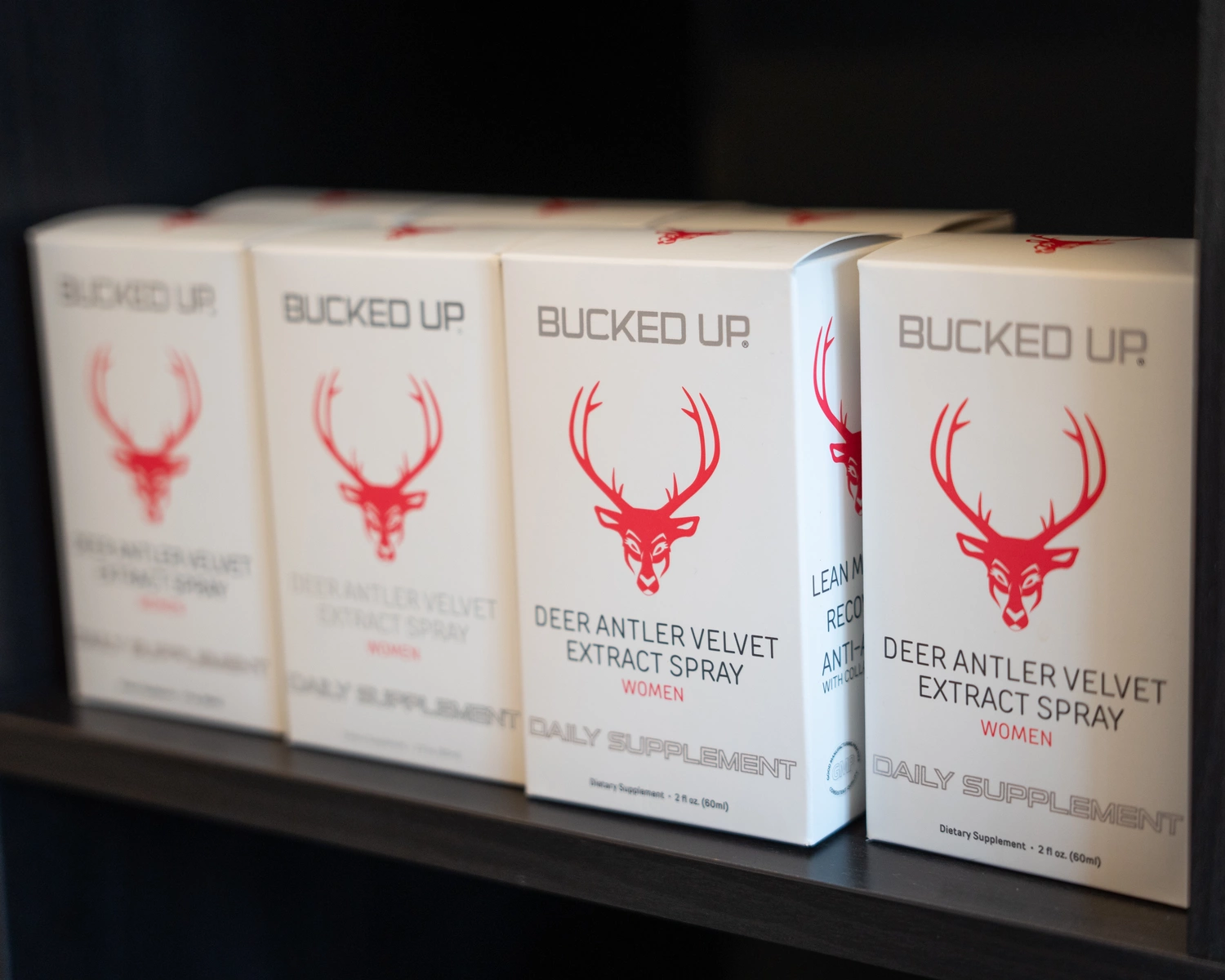A row of white boxes of Bucked Up Deer Antler Velvet Extract Spray for women, featuring a red deer head logo with large antlers, arranged neatly on a black shelf. The boxes are aligned side by side, with the front box slightly more prominent, showing the product name and details. The background is dark, emphasizing the clean, minimalist design of the packaging, with soft lighting highlighting the boxes' surfaces.