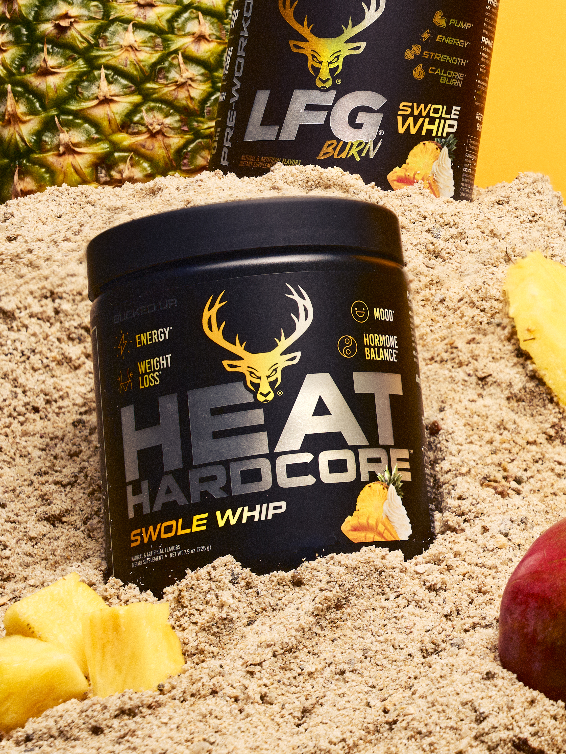 A black container of HEAT HARDCORE Swole Whip dietary supplement with a yellow deer logo and bold gray and yellow text, placed on a sandy surface with pineapple chunks and a red apple nearby, against a bright yellow background, with another similar container partially visible behind it, all arranged in a casual, outdoor setting with natural lighting.